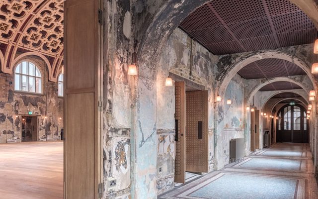 Image of connecting arched side corridor to the Grand Hall. Fire damage shows exposed brick and paint work on the walls leading down to an arched doorway at the end.