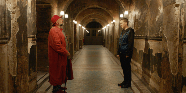 In a corridor with a colourful mosaic floor, Omid Djalili (wearing a long red coat, a red trilby hat, and using a walking stick) faces The Outsider (a person with shaved short hair, wearing all black with a blue vest)