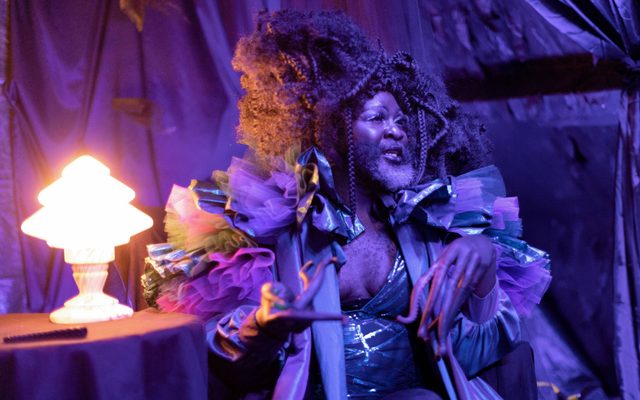 An image of a person with a black and silver beard, large hair decorated with braids sitting next to a lamp on a table. They wear a silver costume with pink and purple ruffled shoulders and have pink tentacles attached to their fingers.