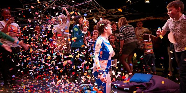Jess Thom, a woman with short brown curly hair wearing a blue and silver superhero suit, is surrounded by children throwing confetti