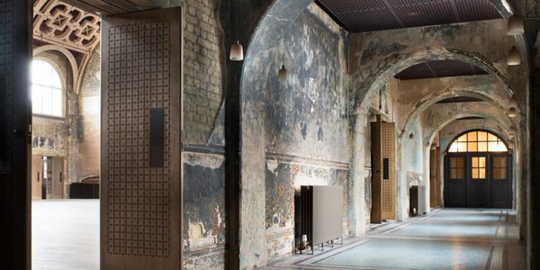A corridor off the Grand Hall, with arches and fire-burnt plaster walls. Through a heavy wooden door on the left hand side, a sliver of the grand hall and the lattice wood ceiling are visible