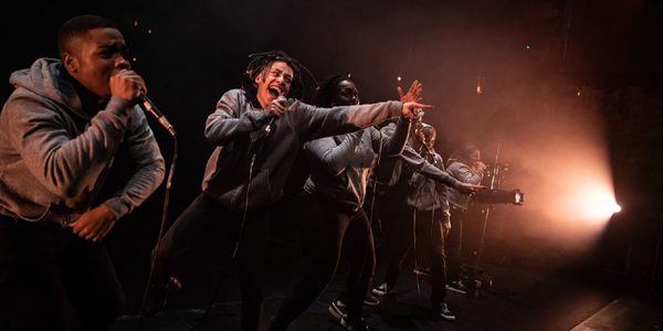 Six beatboxers stand in a row performing into microphones, some pointing outwards, some gesturing with their hands. They all wear grey hoodies and black trousers