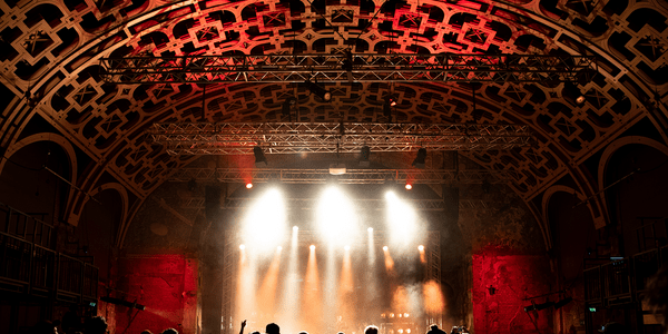 A photo taken from behind a crowd gathered at a concert in our Grand Hall. Spotlights shine down from the wooden lattice roof and illuminate the stage.