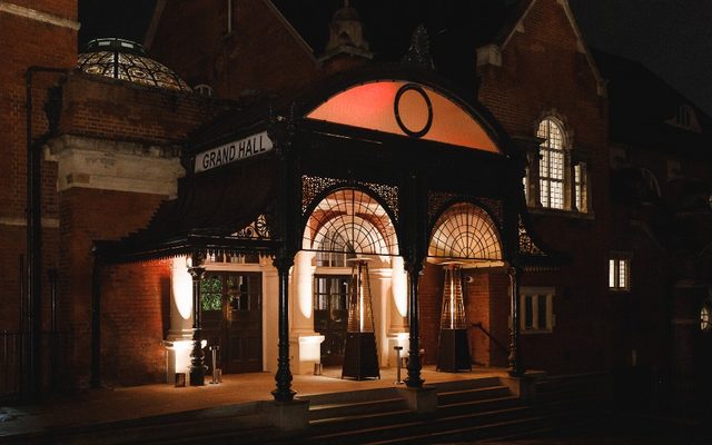 The outdoor entrance to the grand hall, with a wrought iron covering over the steps from the road. It is night and lights are visible from the floor of the entrance platform, a window on the right, and the stain glassed dome on the left.