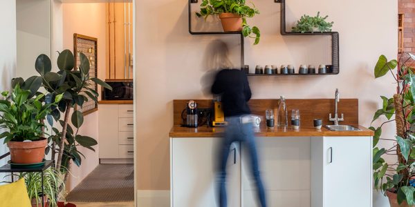 An image of a blurred figure making coffee in the Scratch Hub reception, a single sink unit sits beneath a decorative shelf with large green plants.