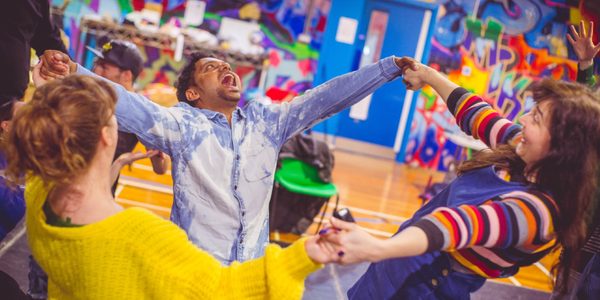 The Happiness Project - three people in brightly coloured clothes hold hands in a circle and are smiling and shouting. The walls of the room are decorated with brightly coloured graffiti