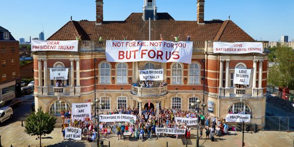 Crowds of people spill out of Battersea Arts Centre's front doors and every balcony. Banners draped across the building read 'Dear President Trump', 'Not For Me, Not For You, But For Us', 'Difference is beautiful', 'Women are powerful', 'More art now', 'Everyone is creative', 'Build bridges not walls', 'United we stand', 'We are open', '#BACPhoenix', 'Love Battersea Arts Centre'.