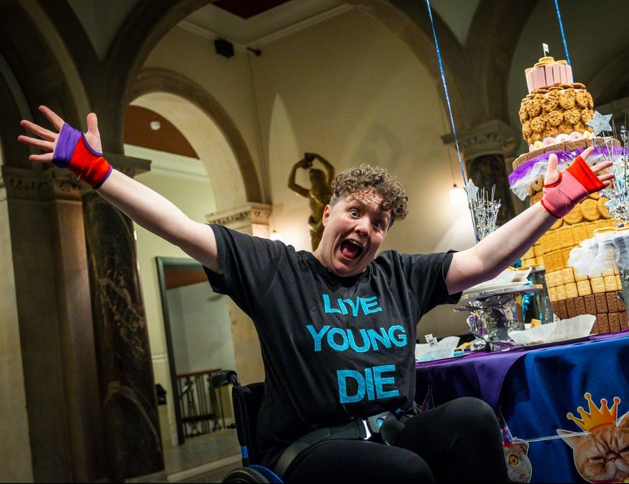 Jess Thom, a woman with short brown curly hair, holds her arms wide in front of a table decorated with cats-in-party-hats bunting and a giant tiered biscuit installation. Photo by Samuel Dore.