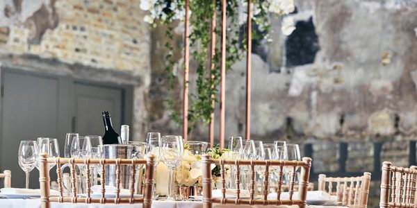 The backs of wooden chairs around a dining table with wine glasses. Ivy leaves hang down from a raised centerpiece. In the background are walls of distressed plaster and exposed bricks.