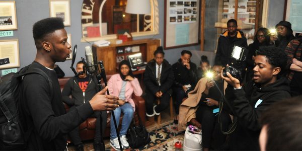 A group of around 15 young people gather in a room, sitting on sofas and chairs. Some are taking photos with their phones. In the foreground one person talks into a microphone with a pop guard, gesturing with his hand, and another points an SLR camera at him