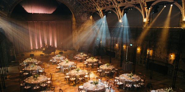 An image of the Grand Hall set up for a wedding. There are lines of circular tables dressed with white table-cloths and flower bouquets. There is a grand piano at the far end of the room and spotlights shining down from the ceiling.