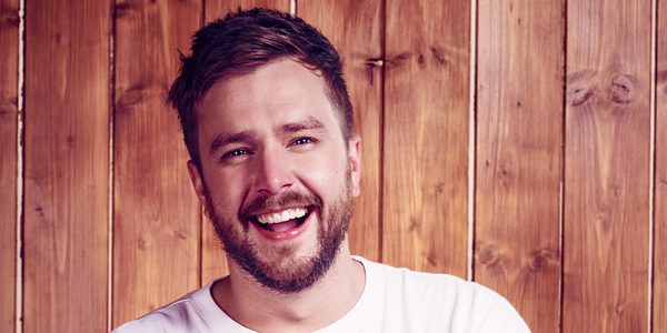 An image of Iain Stirling. He has short brown hair and a short beard wearing a white t-shirt stood against a wood panel backdrop, with a wide smile.