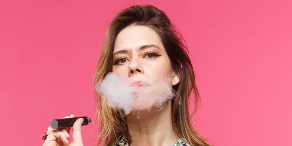 An image of Lou Sanders. A profile from the neck upwards of a women with brown side parted hair looking directly into camera, she holds up a black vape pen in her hand and is releasing smoke from her mouth.