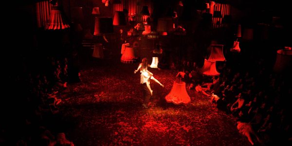 An image from the show Mask of the Red Death. A red hue lights the stage as audience sit cross legged on either side. Light shades hang suspended from the ceiling as a woman dressed in white floats in the centre.