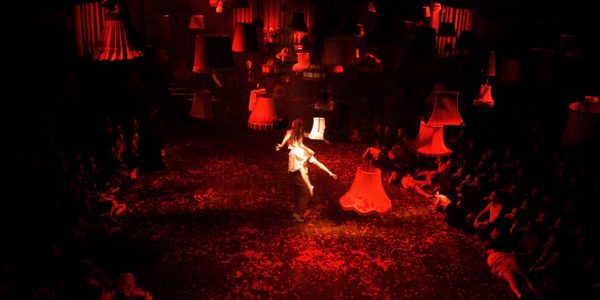 Mask Of The Red Death An image from the show Mask of the Red Death. A red hue lights the stage as audience sit cross legged on either side. Light shades hang suspended from the ceiling as a woman dressed in white floats in the centre.