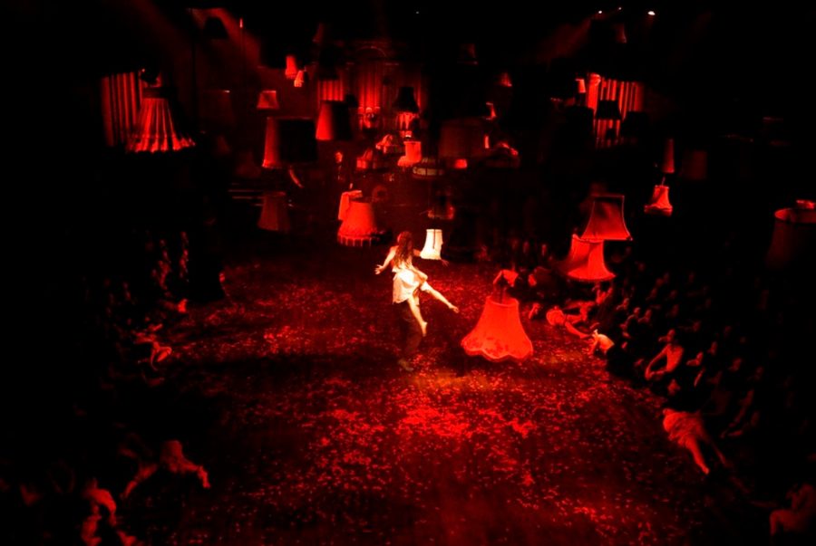 A girl appears to float in the middle of a red lit room, with a number of lampshades also suspended
