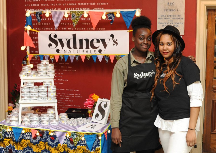 Against a red wall, a stall with colourful bunting is set up for Sydney's Naturals, with lots of branded tins, a colourful tablecloth, and flowers. Sydney, wearing a black branded apron and green shirt, and another agent, wearing a black fedora, branded black t shirt, and white trousers, stand to the right of the stall