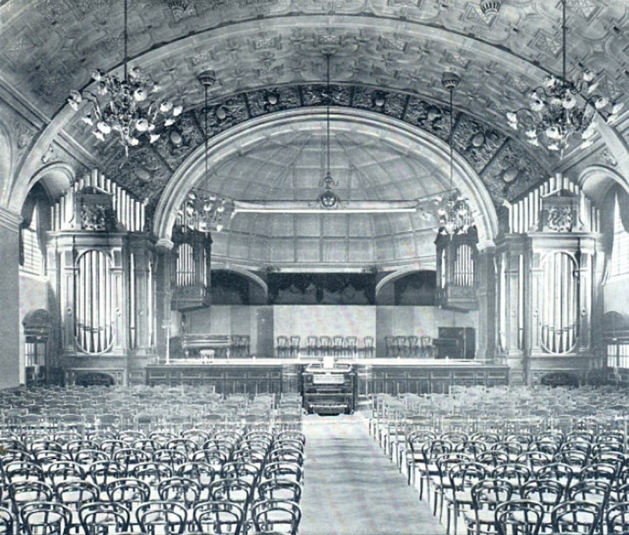 A black and white image of the grand hall, with chandeliers hanging from the ceiling, organ pipes on each side of the stage, the organ console in the middle in front of the stage, and chairs laid out all through the hall with an aisle in the middle