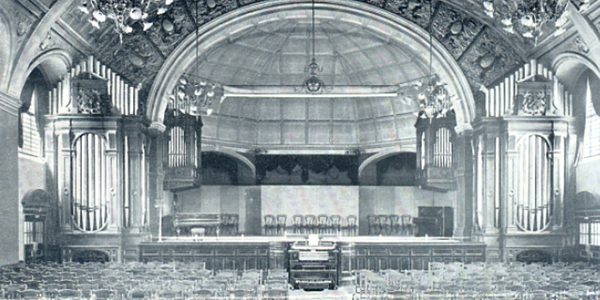A black and white image of the grand hall, with chandeliers hanging from the ceiling, organ pipes on each side of the stage, the organ console in the middle in front of the stage, and chairs laid out all through the hall with an aisle in the middle