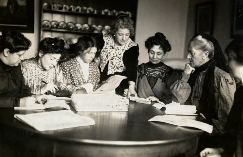 A black and white photograph of seven women around a table, with open books and papers. Six are sitting, one stands in the middle pointing at something in one of the books