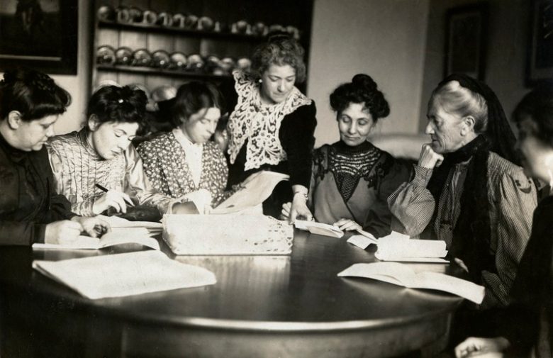 Suffragetes A black and white photograph of seven women around a table, with open books and papers. Six are sitting, one stands in the middle pointing at something in one of the books