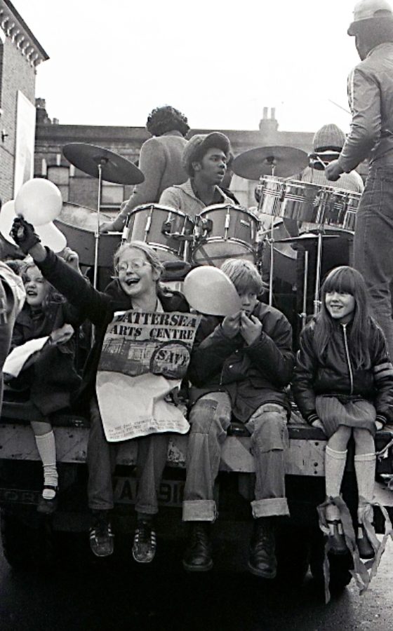 A black and white photograph of a young man playing a drum kit, while in front of him four children sit with their legs hanign off the stage, holding balloons. One girl with glasses holds a poster which says 'Battersea Arts Centre' at the top