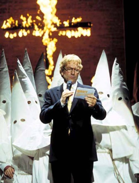 A still from 'Jerry Springer The Opera'. A cross burns in the background as a man with blonde hair and glasses in a suit stands in front of a group of people in white pointy hoods and robes. The man holds a microphone and Jerry Springer cue cards.