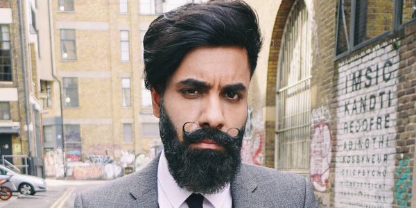 An image of Paul Chowdhry. A dark haired man wearing a suit. His hair is quaffed with a long beard and curled moustache. He looks directly into the camera with a backdrop of a city alleyway.
