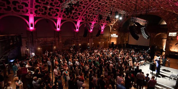 Proms at Battersea Arts Centre_Hires_16.9 A large standing crowd in the Grand Hall, with the ceiling lit up pink. A stage in the middle of the right hand side has a musician and a harp. Projected onto the far wall are logos for BBC Radio and BBC Proms