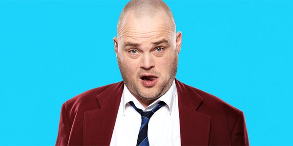 An image of Al Murray, a man with a shaved head wearing a maroon blazer, white shirt, blue & navy striped tie and a belt buckle with the word 'GUV' on it. He stands in front of a light blue background looking directly into camera.