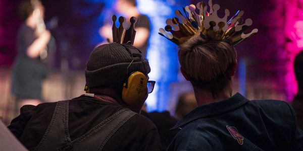 The back silhouette of two figures one wearing ear defenders and the other wearing several paper crowns.