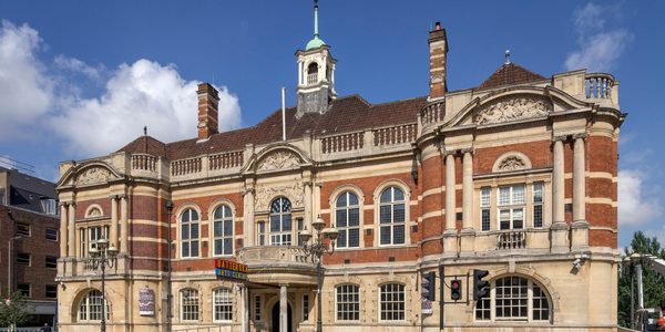 The front of our building, a Victorian town hall, with pale stone on the ground floor, red bricks on the first floor with pale columns and outlines on the windows, and a balcony around the roof. Our red, blue and yellow sign is at the front. Bollards and lamposts separate the pavement from the road. In the background the sky is blue with a few clouds.