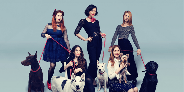 A group of women against a pale blue background. Three women stand and two crouch next to 5 black and white dogs.