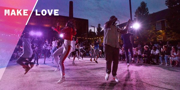 An image of a group of young people dancing and leaping in a circle surrounded by an audience in an industrial estate.