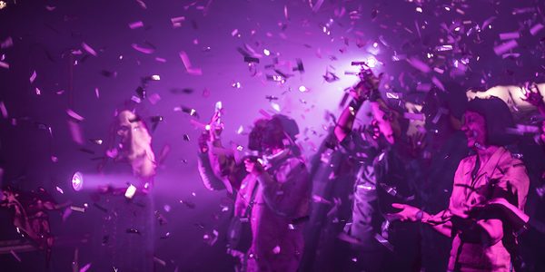 Hazy, purple lights wash over a group of people, who stand, arms outstretched, amid a burst of purple confetti.