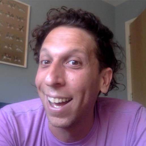 A white man with curly brown hair and a pink t-shirt smiles into the camera.