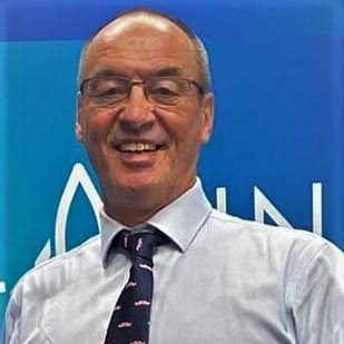 A middle aged white man in a shirt, tie and glasses is smiling. Behind him is a blue background.