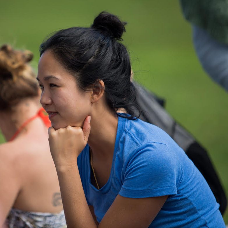 An Asian woman in profile wearing a blue t-shirt and her hair in a top knot. Her chin rests on her left fist.