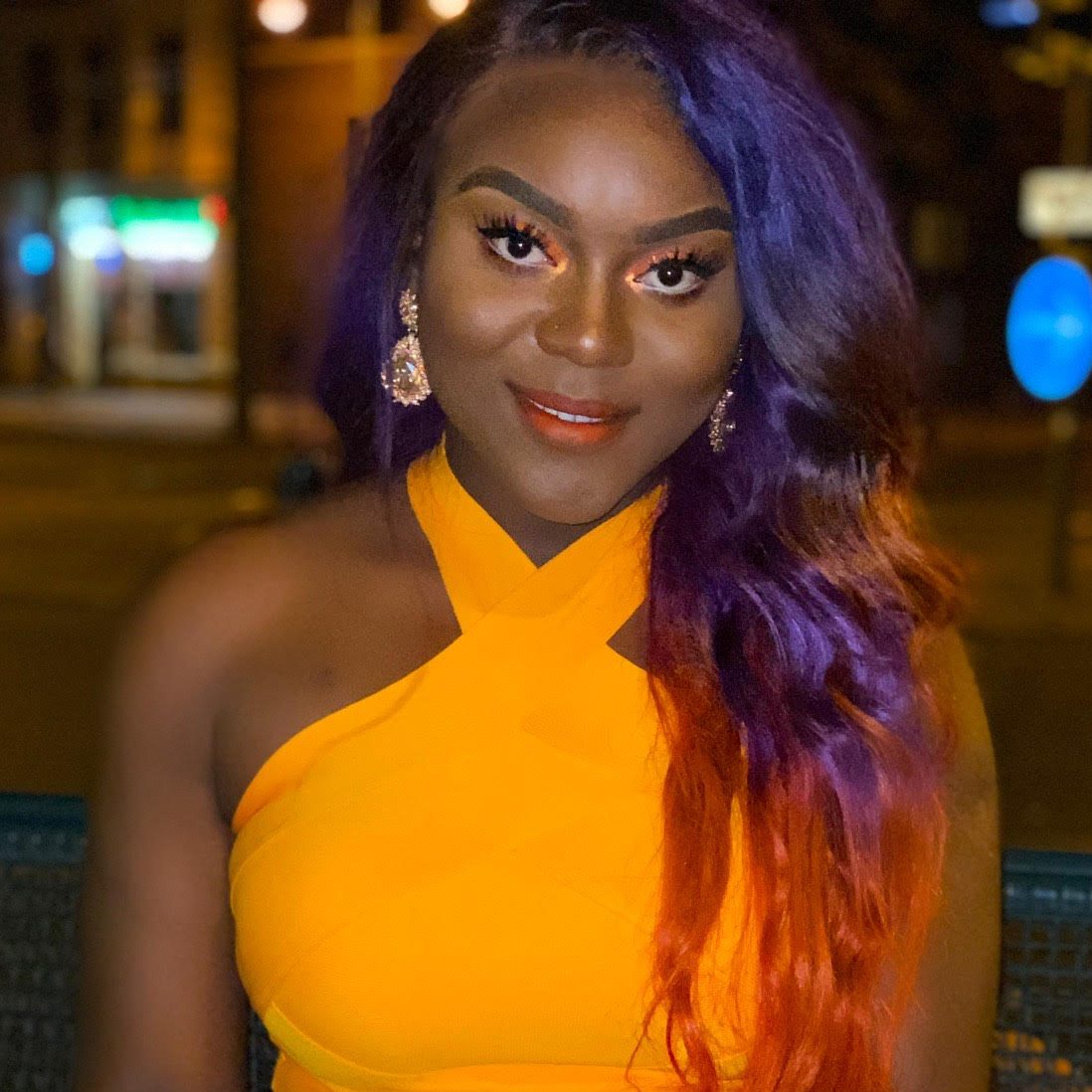 A young black woman with long purple hair and a bright orange halterneck dress.