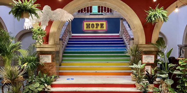 An image of the Battersea Arts Centre foyer. An alcove entrance over a grand rainbow stair case. To either side, there are plants hanging from the ceiling and gathered in pots on the floor. A yellow and pink sign saying 'HOPE' can be seen at the top of the steps.
