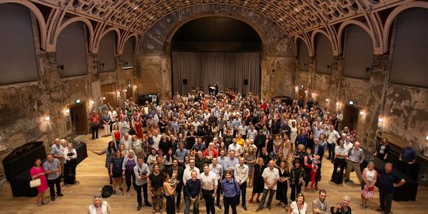 A crowd of people fill the grand hall. There are lights in the wood pannelled ceiling and the back of the hall is covered with a grey curtain