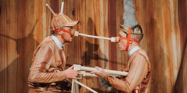 Two people wearing costumes that look like wood are standing in front of a wood-effect backdrop. The have wooden poles attached to their noses. They are facing each other and the poles on their noses are touching.