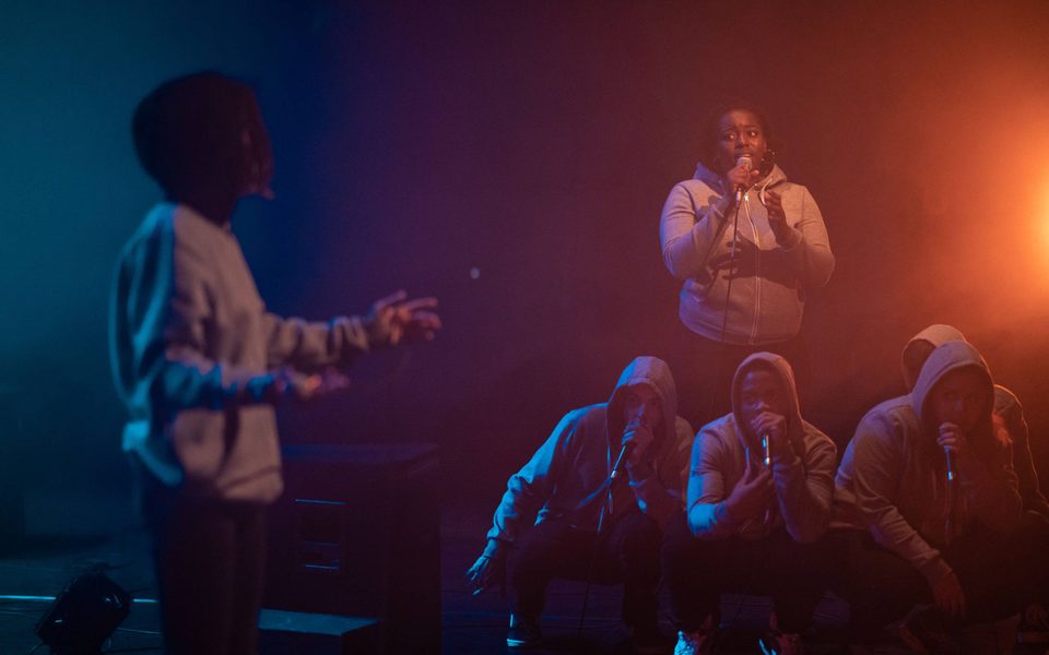 4 beatbox performers huddle together looking back at a solo performer, blurred at the front of the shot