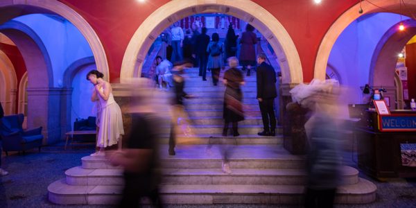 The front staircase in the foyer with red walls and blue lighting. People are walking up the stairs as dancers stand and move around them.