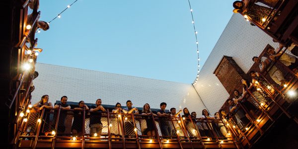 A view looking up at the corner of the standing gallery, with audience members looking over onto the stage, in our courtyard, draped in lightbulbs