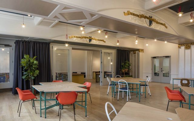 The Lower Hall: a large space with woodens floors, white walls, groups of tables surrounded by chairs. There are large plants, dark blue curtains and glass doors leading to another space.