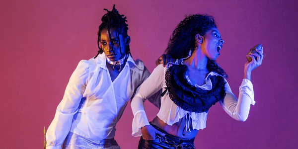 Two dancers strike a pose, one is going to bite a pear that they are holding. They are lit by a blue light against a purple background.