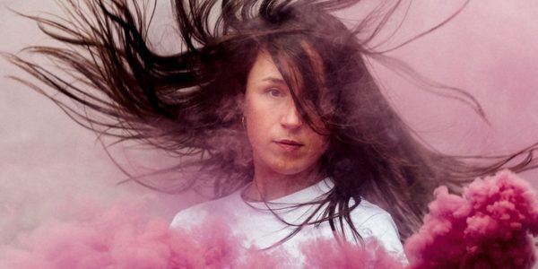 A woman wearing a white t-shirt with long brown hair blowing; holding a pink smoke bomb with pink smoke billowing out.