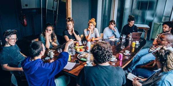 Scene Change 2022 image A group of people are sat around a large wooden table. One person is speaking and other people are looking at them.