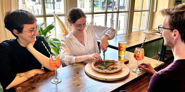 Three people sat around a table in the bar; they have drinks in front of them and one person is cutting a pizza.